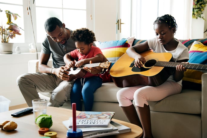 Black father on the couch with two young daughters who both have guitars as he teaches them to play guitar