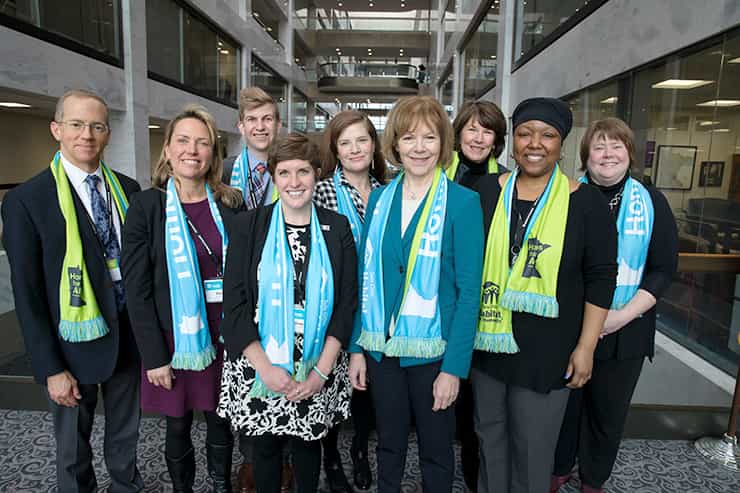 Advocates from Twin Cities Habitat for Humanity and other MN Habitat affiliates met with MN Senator Tina Smith (fourth from right) at Habitat on the Hill D.C. in 2019. They are standing in an office building atrium and are all wearing light blue and green Habitat scarves.