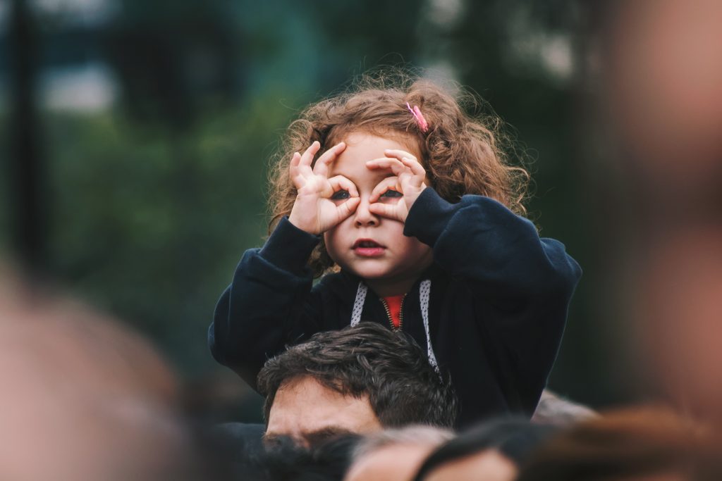 O que é psicomotricidade infantil e como promovê-la? 7 Criança menina sentada nos ombros dos pais com as mãos nos olhos, representando a psicomotricidade infantil.