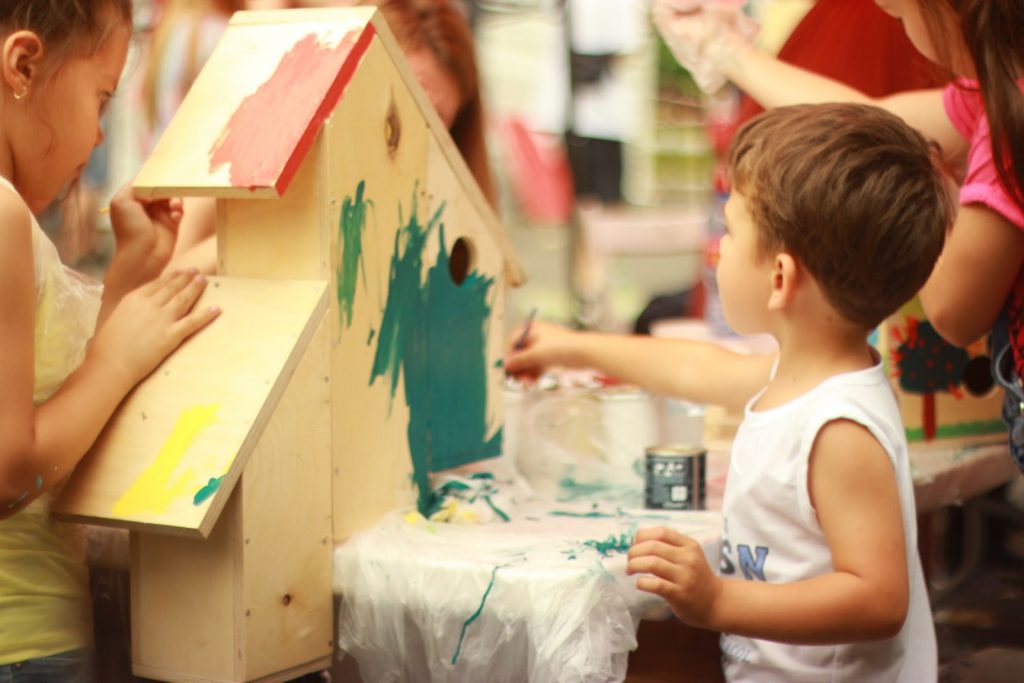 Duas crianças se reúnem para pintar uma casinha de madeira, na imagem, em uma atividade em família para exercitar a criatividade infantil.