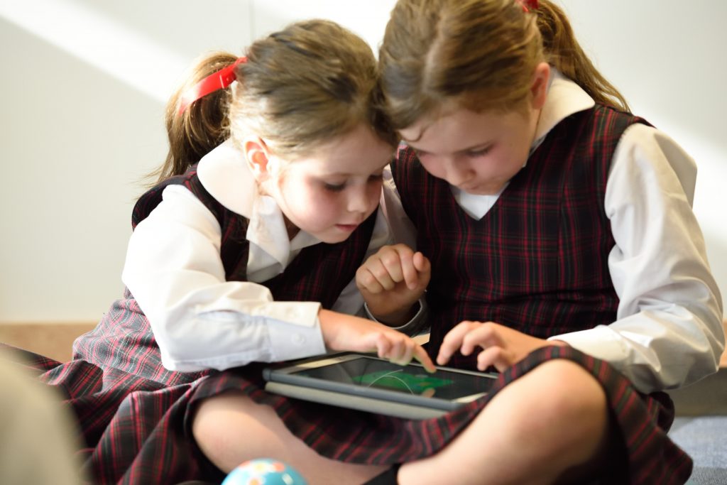 Importância do inglês nos primeiros estágios da vida 1 Duas meninas crianças com uniformes escolares brincando com um tablet demonstrando a importância do inglês.