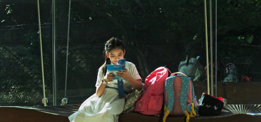 O que é aluno protagonista? 1 Uma menina exemplificando o que é aluno protagonista, lendo um livro ao lado de mochilas.
